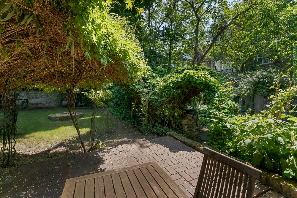Garten mit Terrasse, Holztisch, Stuhl und von Pflanzen überwuchertem Torbogen.