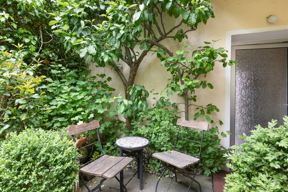 Kleiner Gartenbereich mit zwei Holzstühlen, rundem Tisch, viel Grün und einer Tür im Hintergrund.