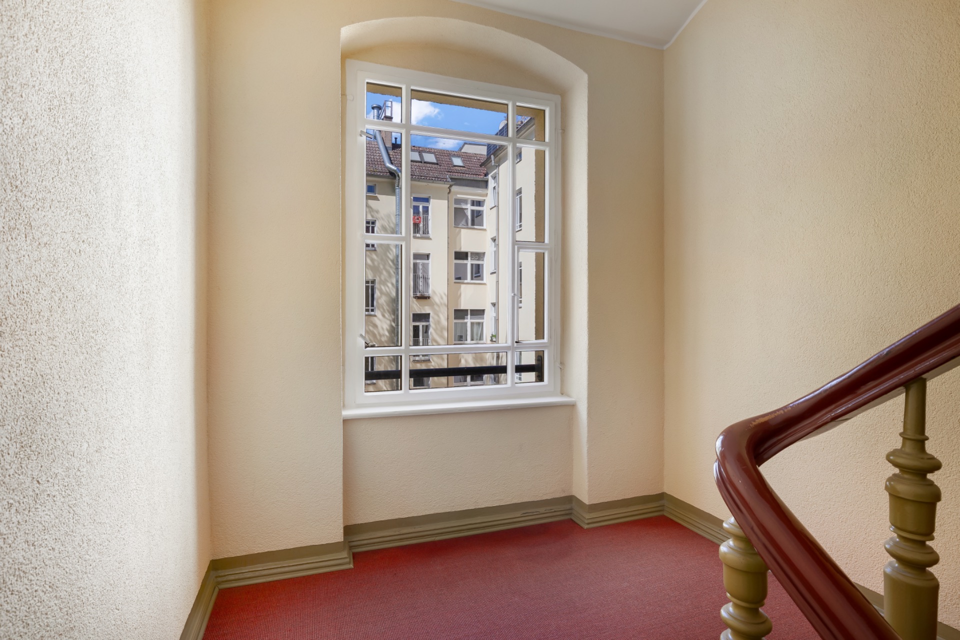 Treppenhaus mit Fenster und rotem Teppichboden, Blick auf Nachbargebäude.