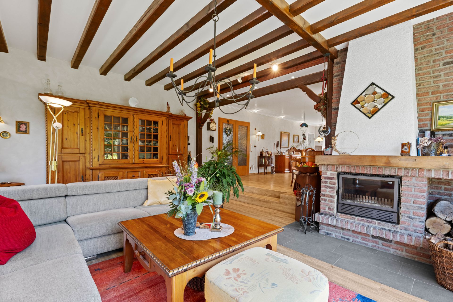 Helles Wohnzimmer mit Holzbalkendecke, Kamin, Sofa und Holztisch mit Blumenstrauß.