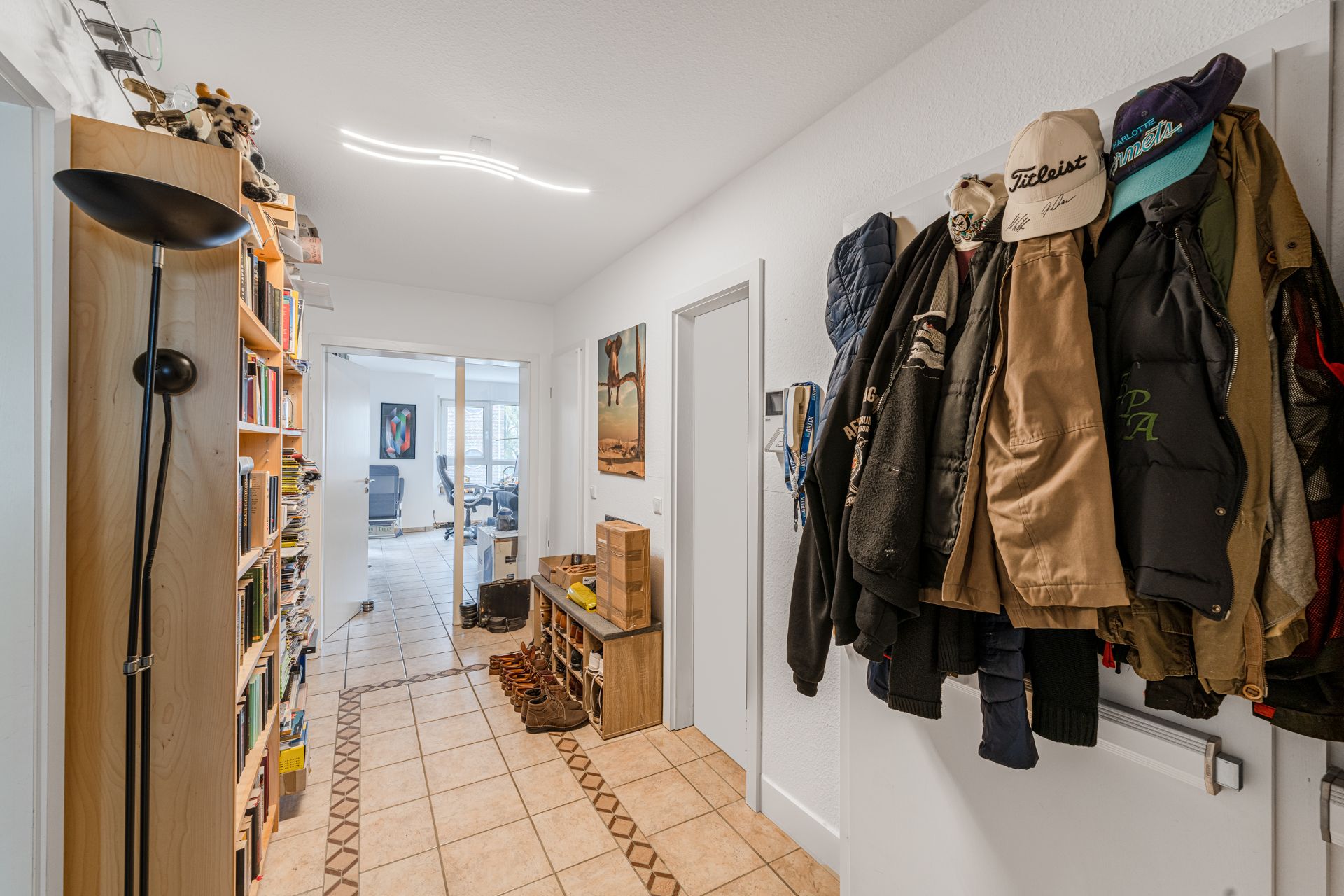 Flur mit Bücherregal, Garderobe voller Jacken und Blick ins helle Zimmer mit Schreibtisch.