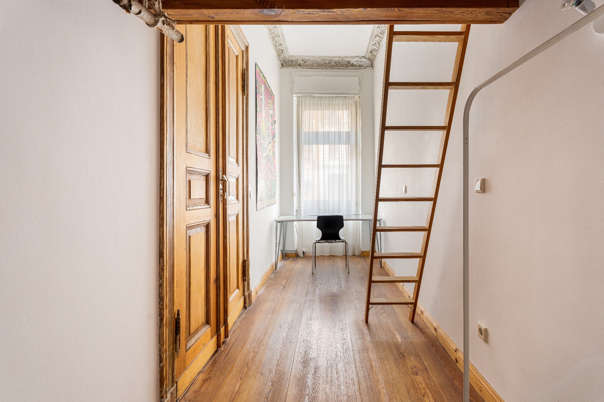 Schmaler Raum mit Holzboden, Leiter, Fenster, Schreibtisch und schwarzem Stuhl.