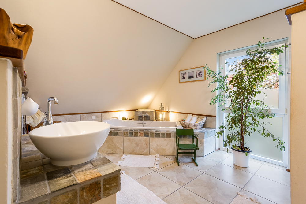 Helles Badezimmer mit Eckeckbadewanne, großem Waschbecken und großer Zimmerpflanze am Fenster.