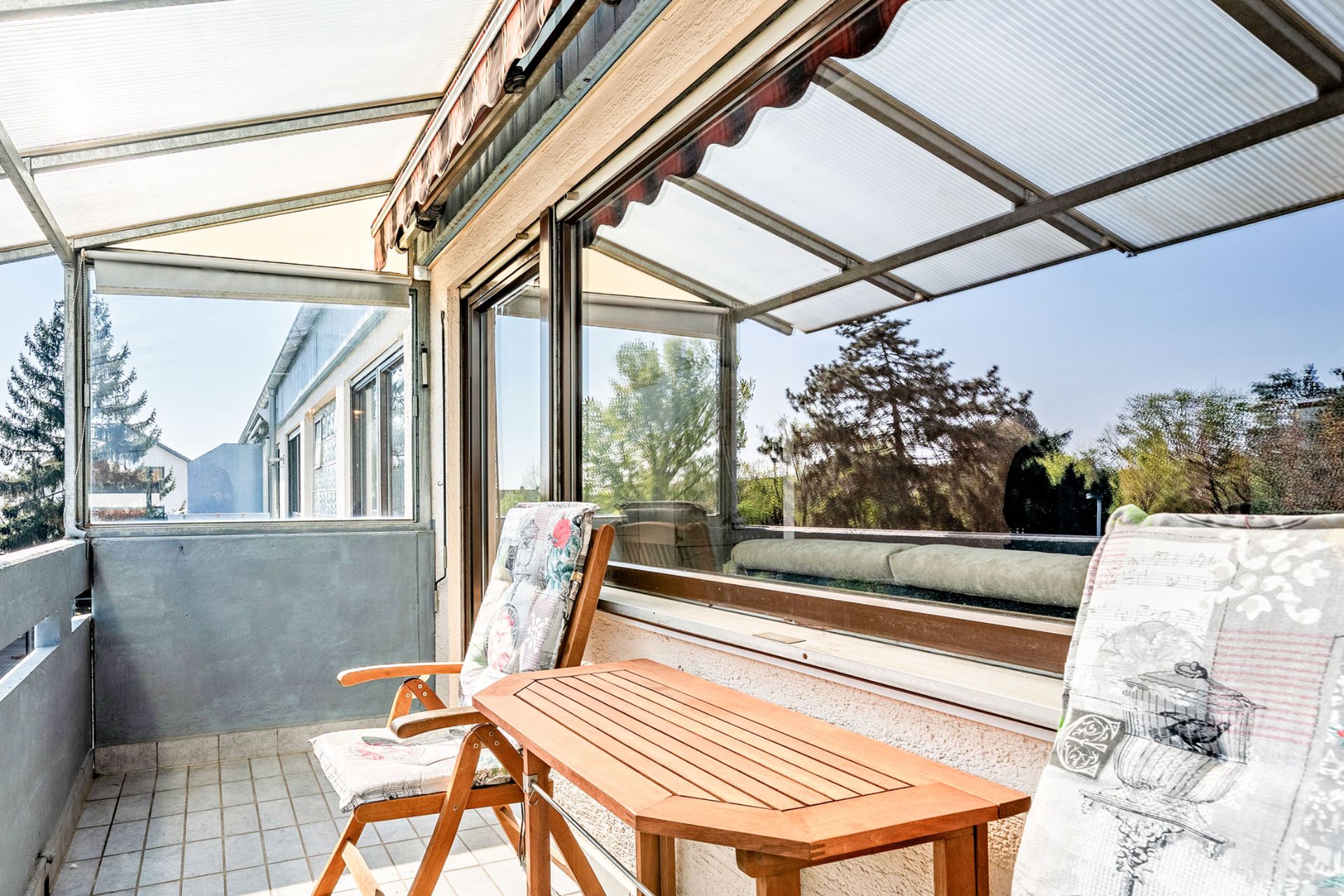 Überdachter Balkon mit zwei Stühlen und einem Holztisch, Blick auf Bäume im Hintergrund.