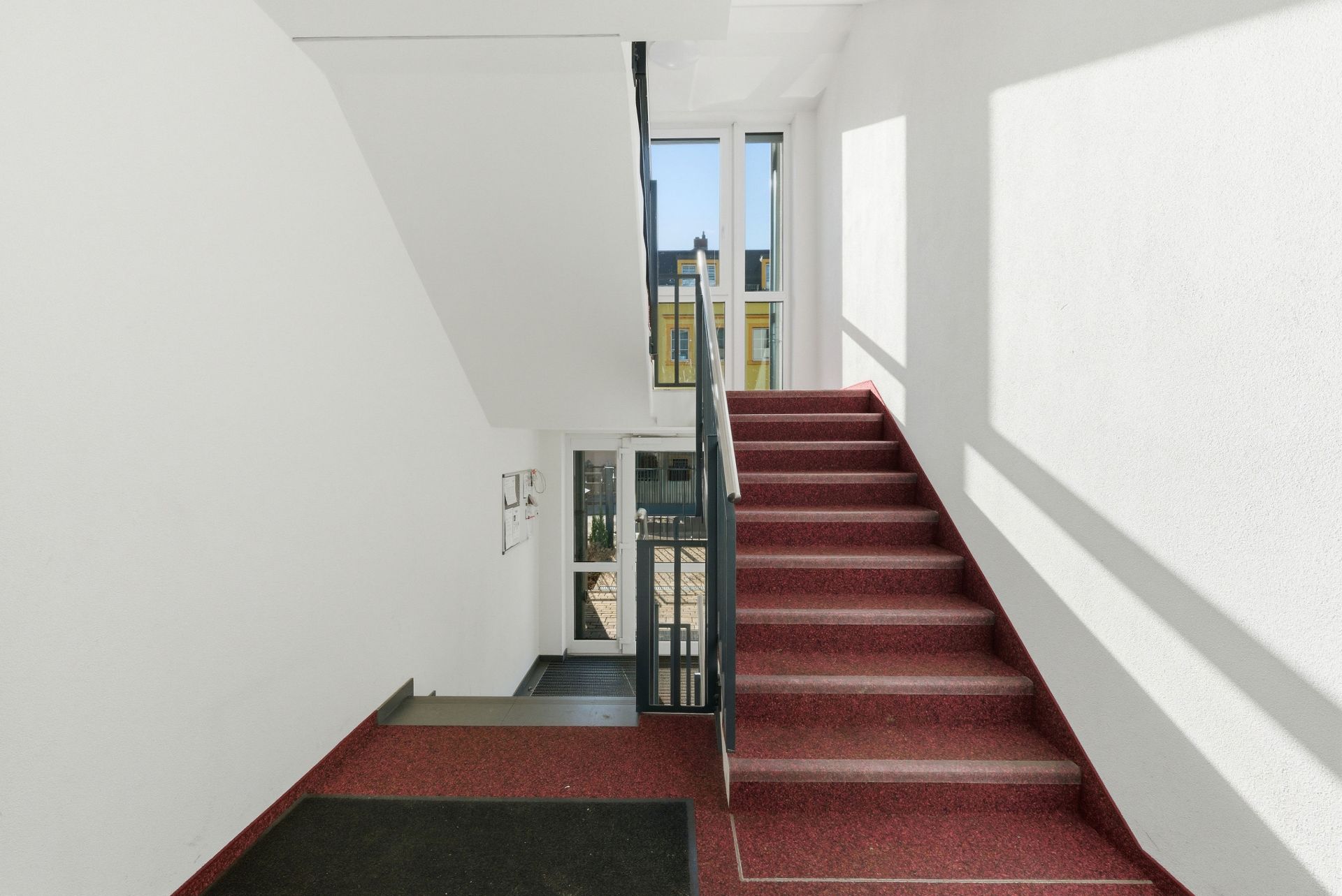 Helles Treppenhaus mit roten Stufen und großen Fenstern in einem Wohnhaus.