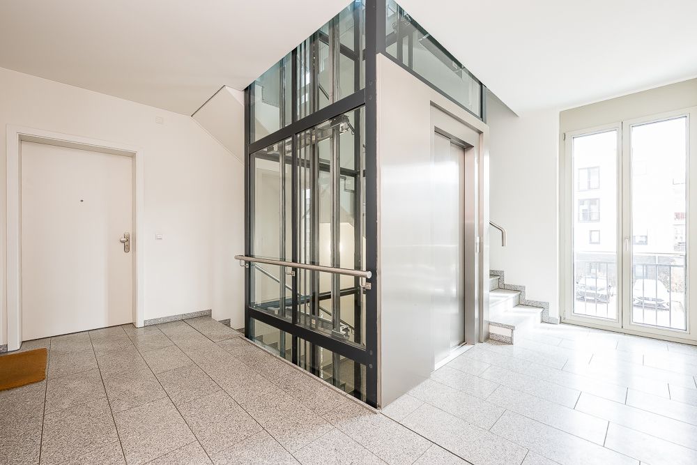 Heller Flur mit Aufzug, Treppe und einer Wohnungstür in einem Wohngebäude.