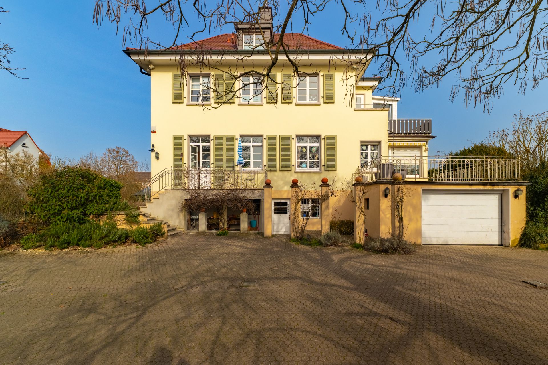 Großes gelbes Wohnhaus mit grünlichen Fensterläden, Garage und gepflastertem Hof.