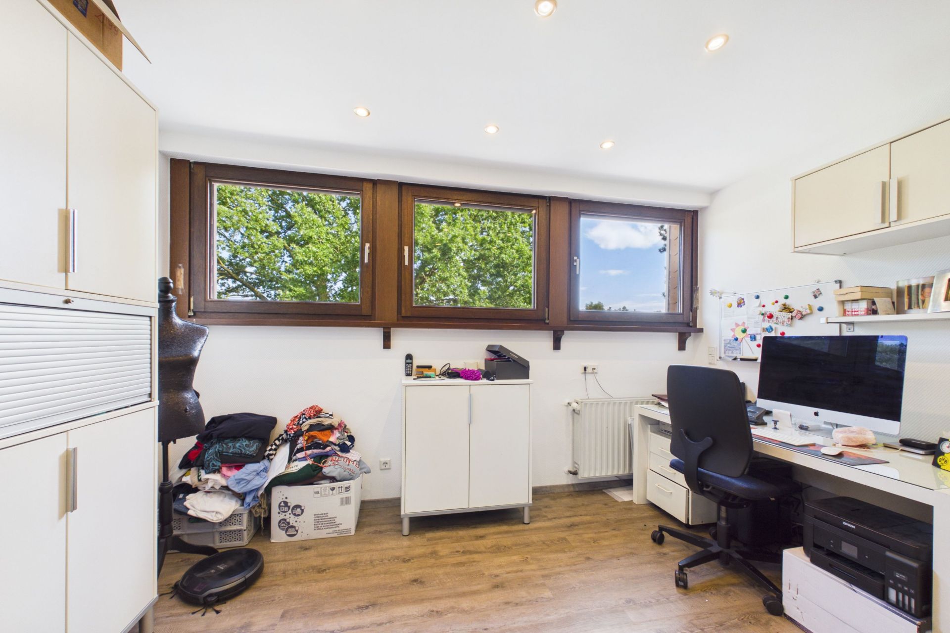 Helles Arbeitszimmer mit Schreibtisch, Computer, Schränken und großen Fenstern mit Blick ins Grüne.