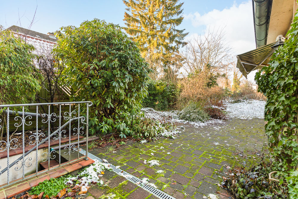Gepflasterte Terrasse mit etwas Schnee, Büschen, einem Baum und einem Geländer im Vordergrund.