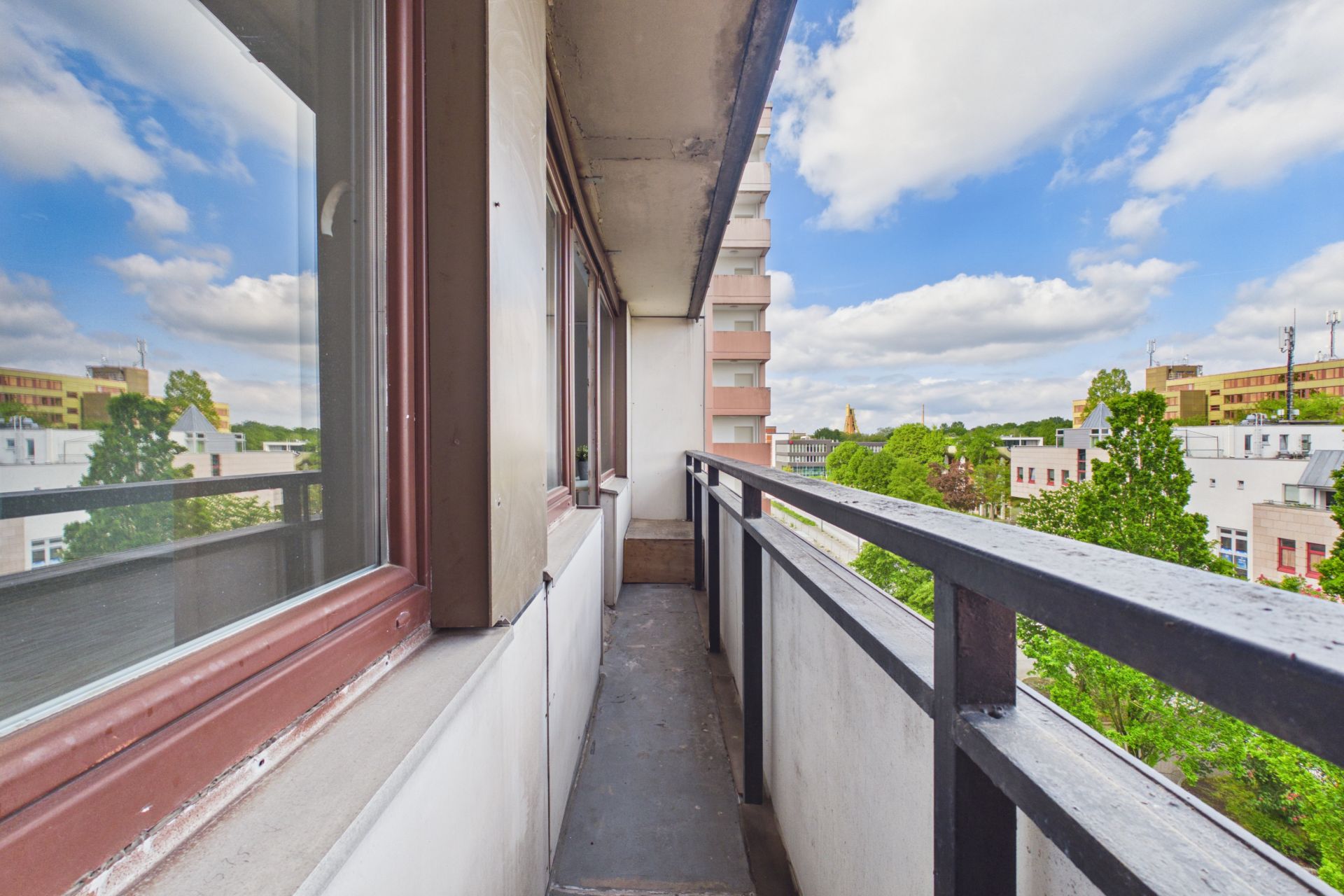 Schmaler Balkon mit Geländer, Blick auf Bäume und Gebäude unter blauem Himmel mit Wolken.