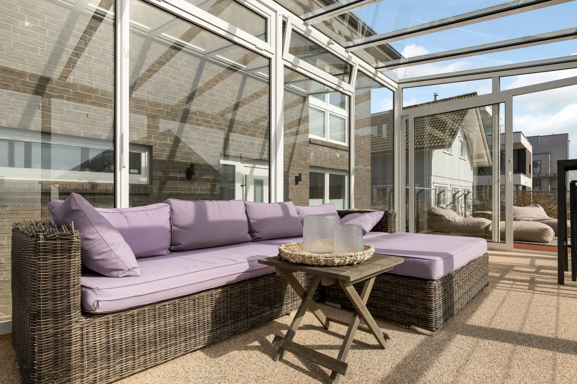 Wintergarten mit Rattansofa, lila Kissen und kleinem Holztisch mit Deko auf hellem Boden.