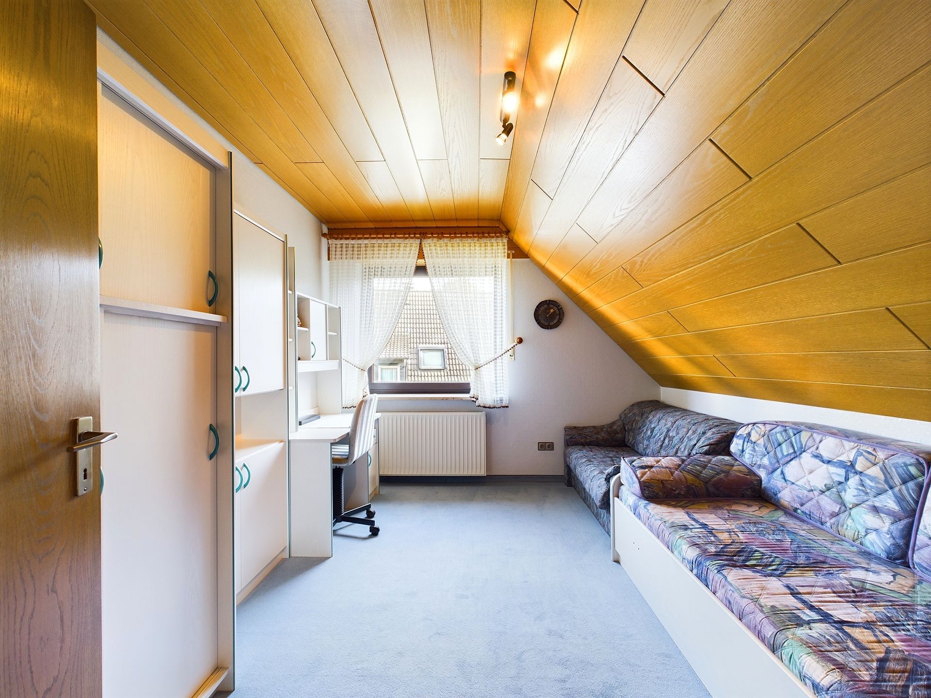 Helles Zimmer mit Holzdecke, Sofa, Schreibtisch und Fenster mit Gardine.