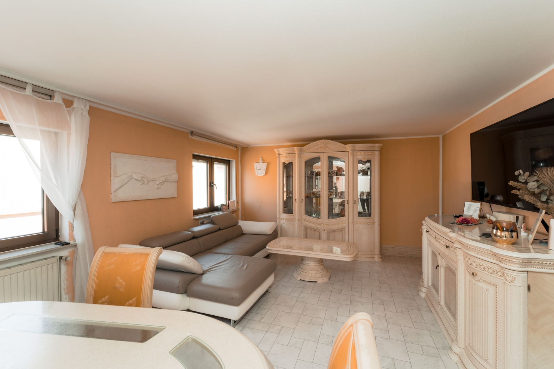 Wohnzimmer mit Ecksofa, Glaskommode, Esstisch und großem Fernseher an der Wand.