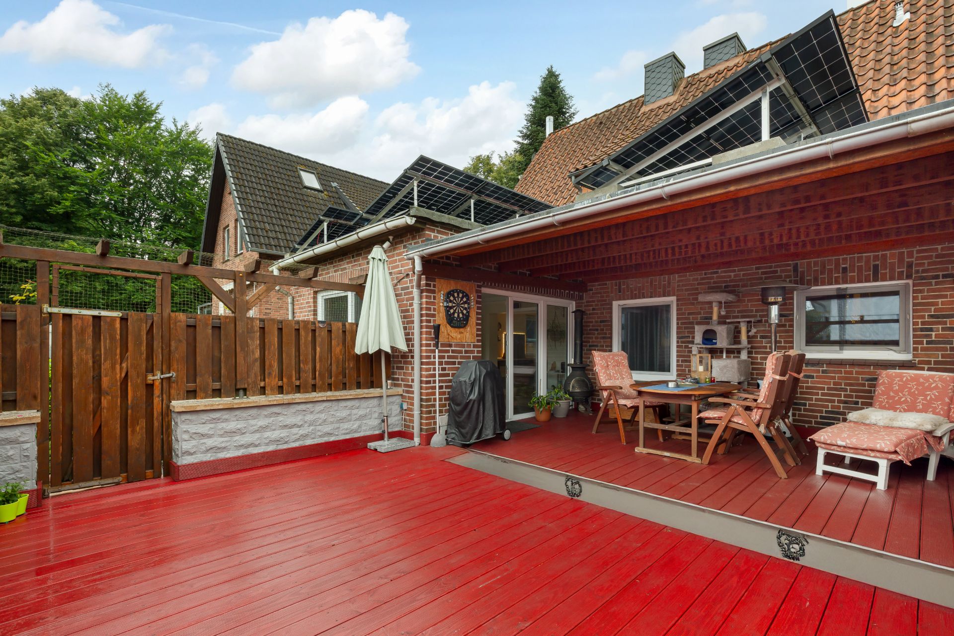Überdachte Terrasse mit Holztisch, Stühlen und rotem Holzboden neben einem Wohnhaus.