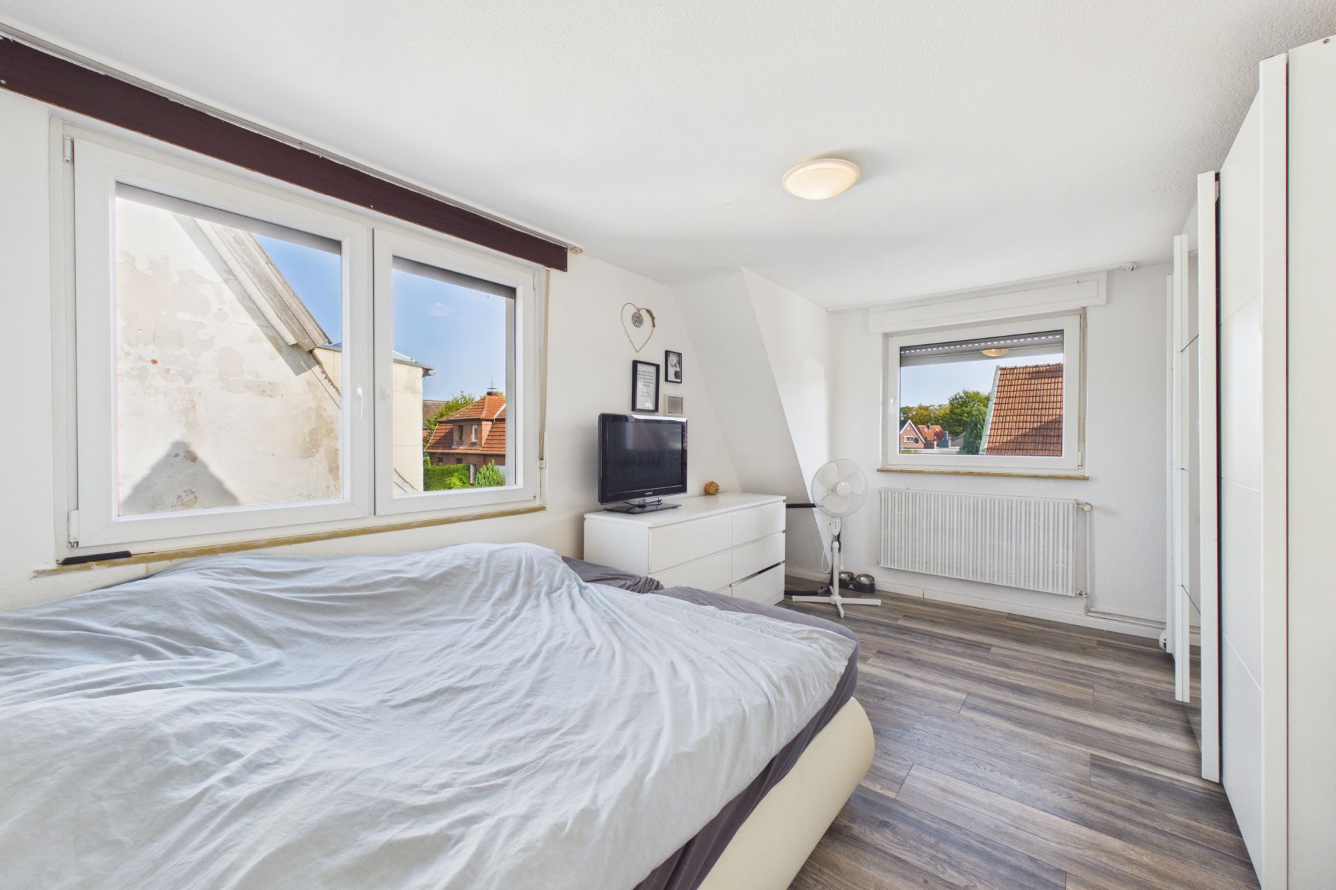 Helles Schlafzimmer mit Doppelbett, Kommode, Fernseher und zwei Fenstern mit Blick nach draußen.