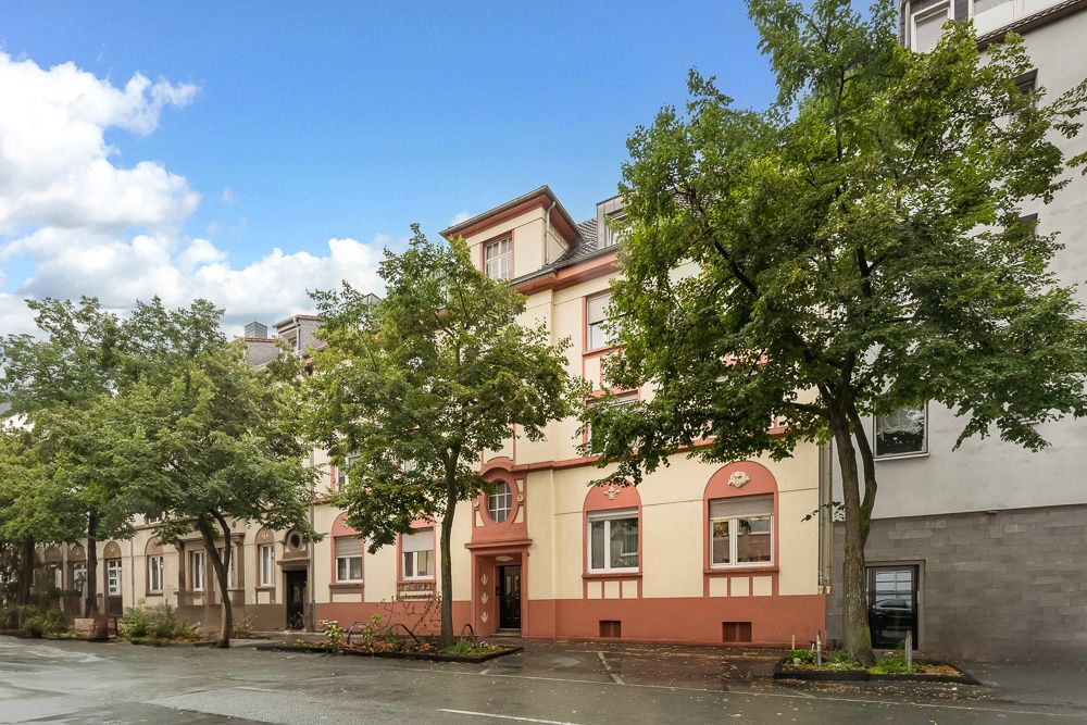 Mehrstöckiges Wohngebäude mit beigen und roten Fassaden, Bäumen und nasser Straße im Vordergrund.