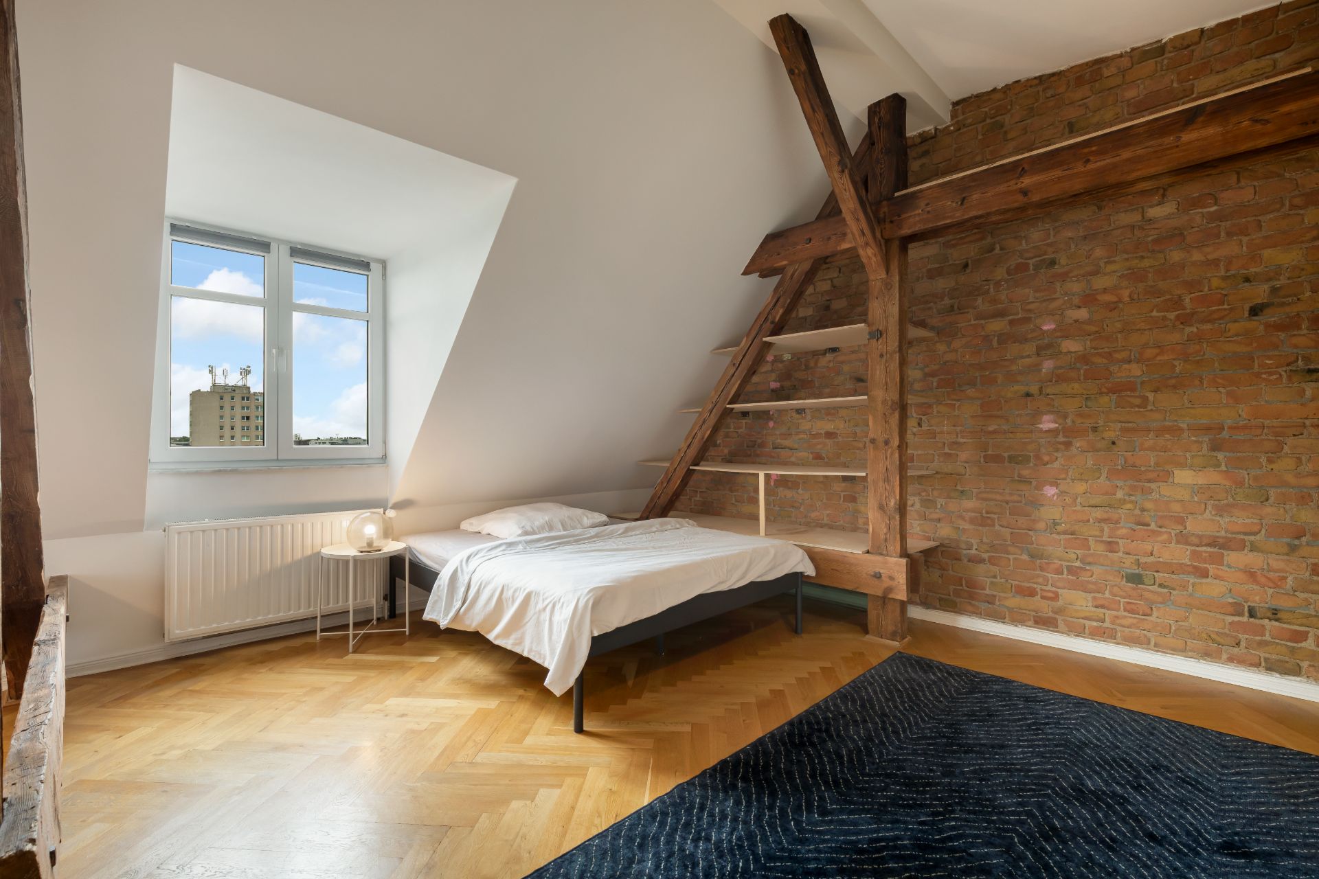 Helles Schlafzimmer mit Holzboden, Backsteinwand, Bett, Fenster und Holzregalen unter Dachschräge.