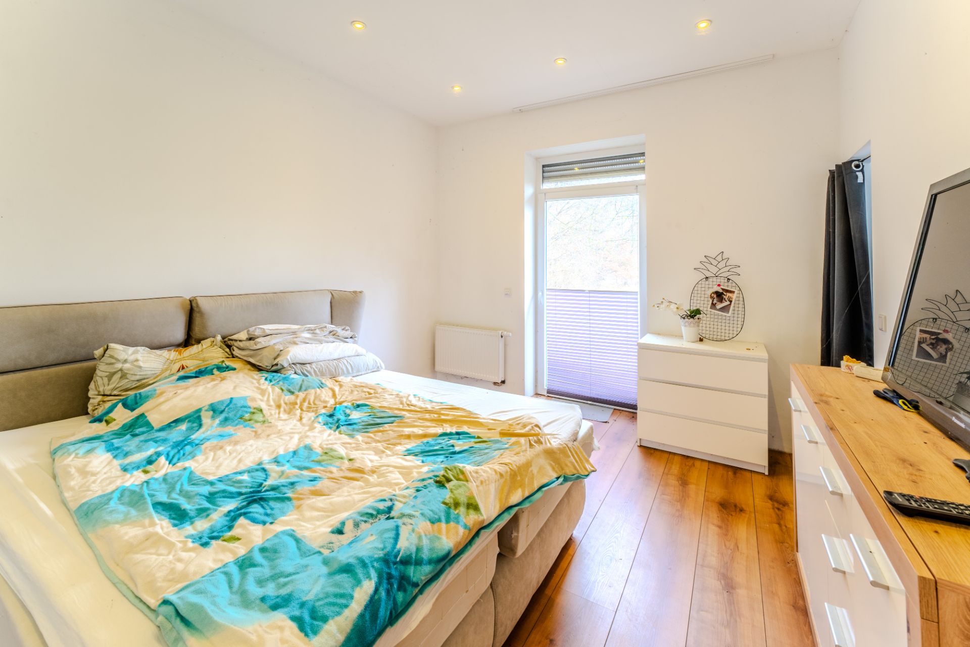 Schlafzimmer mit ungemachtem Bett, Holzboden, Kommode und Fenster mit Jalousie.