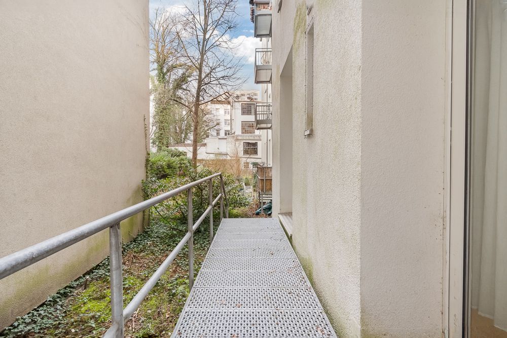 Metallrampe mit Geländer zwischen zwei Gebäuden, Blick auf Innenhof und Bäume.