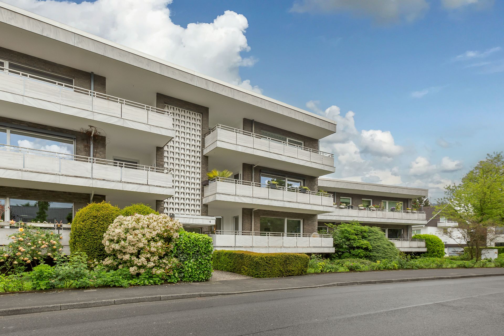 Modernes Wohnhaus mit mehreren Balkonen und viel Grün vor dem Gebäude.