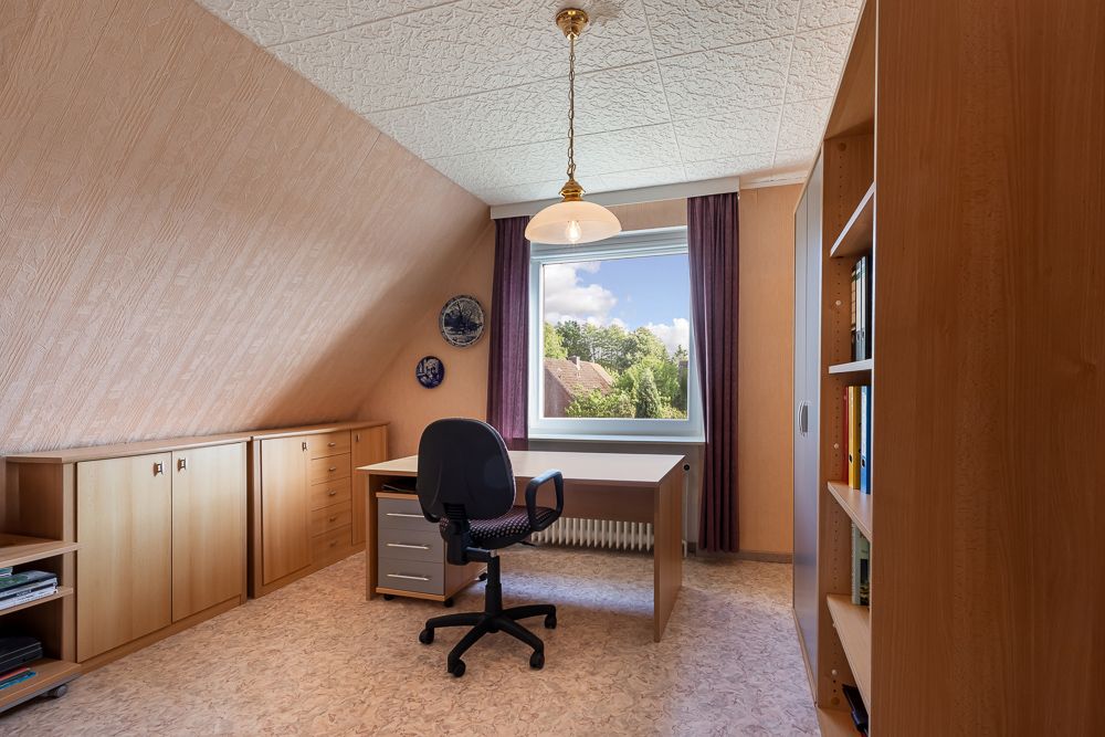 Helles Arbeitszimmer mit Schreibtisch, Stuhl, Regal und großem Fenster mit Vorhängen.