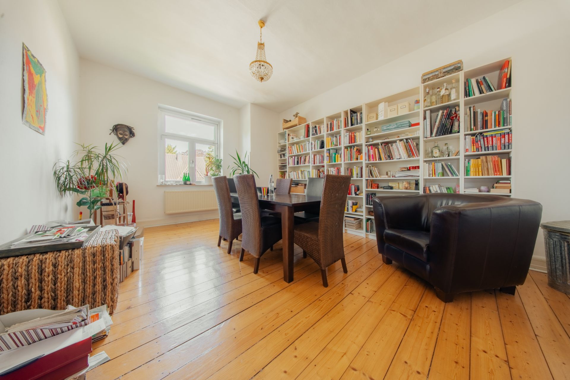 Heller Raum mit Holzboden, großem Bücherregal, Esstisch und Stühlen, Sessel und Pflanzen.
