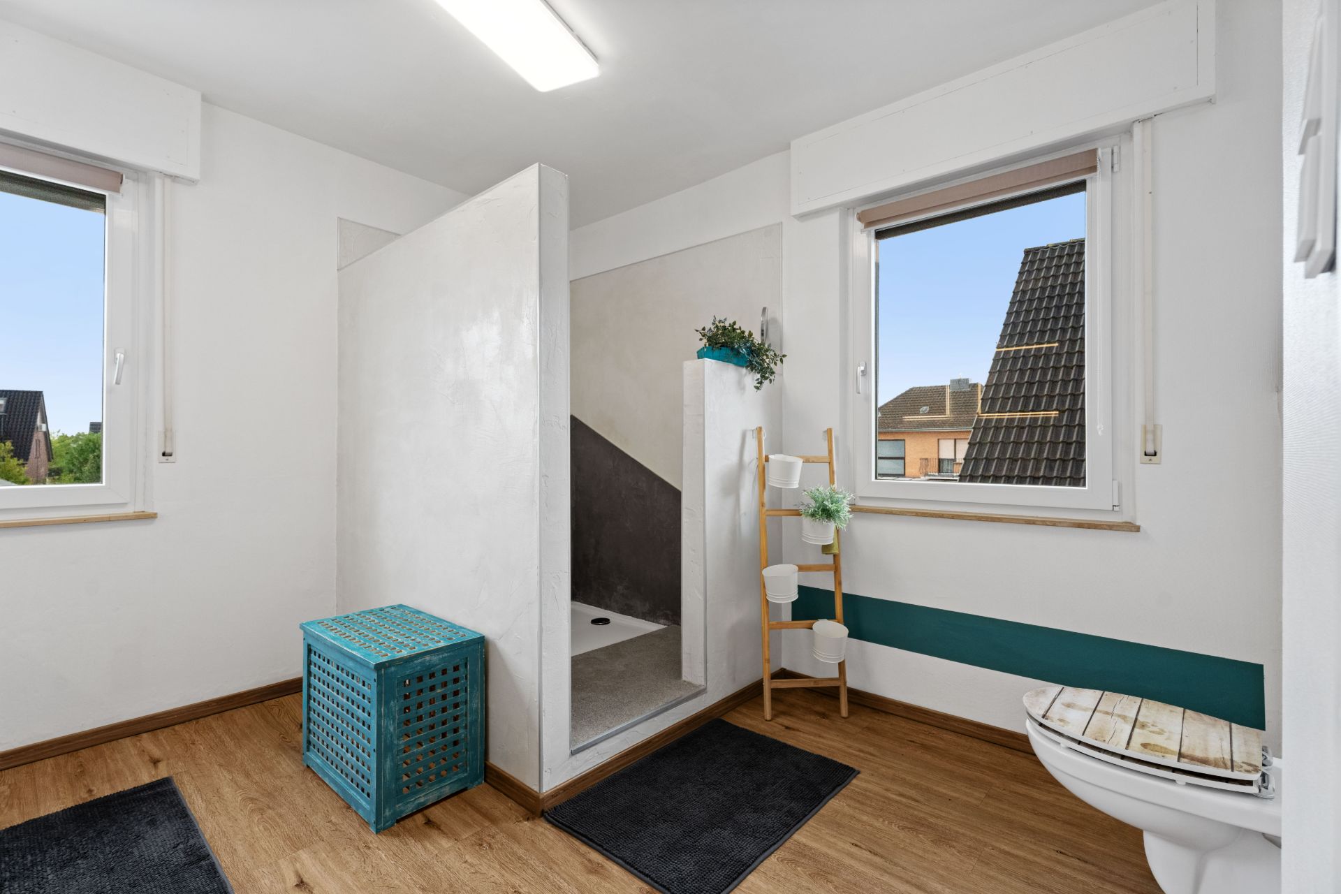 Helles Badezimmer mit Dusche, zwei Fenstern, WC, Holzdetails und einem blauen Wäschekorb.