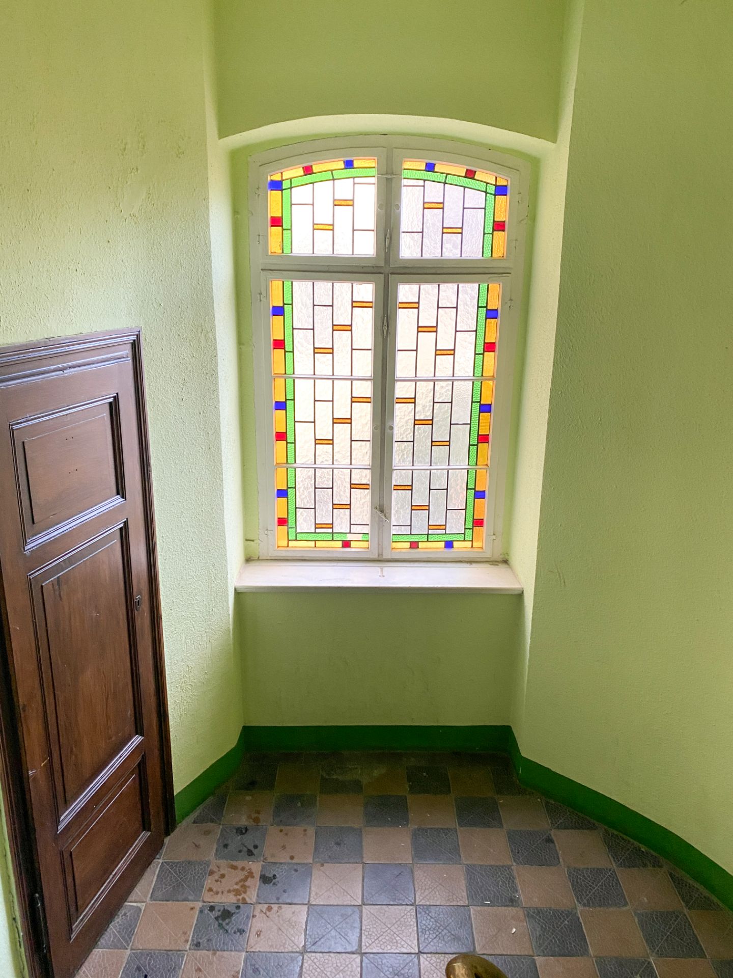 Fenster mit buntem Glas, daneben braune Holztür, Fliesenboden in einem grünen Treppenhaus.