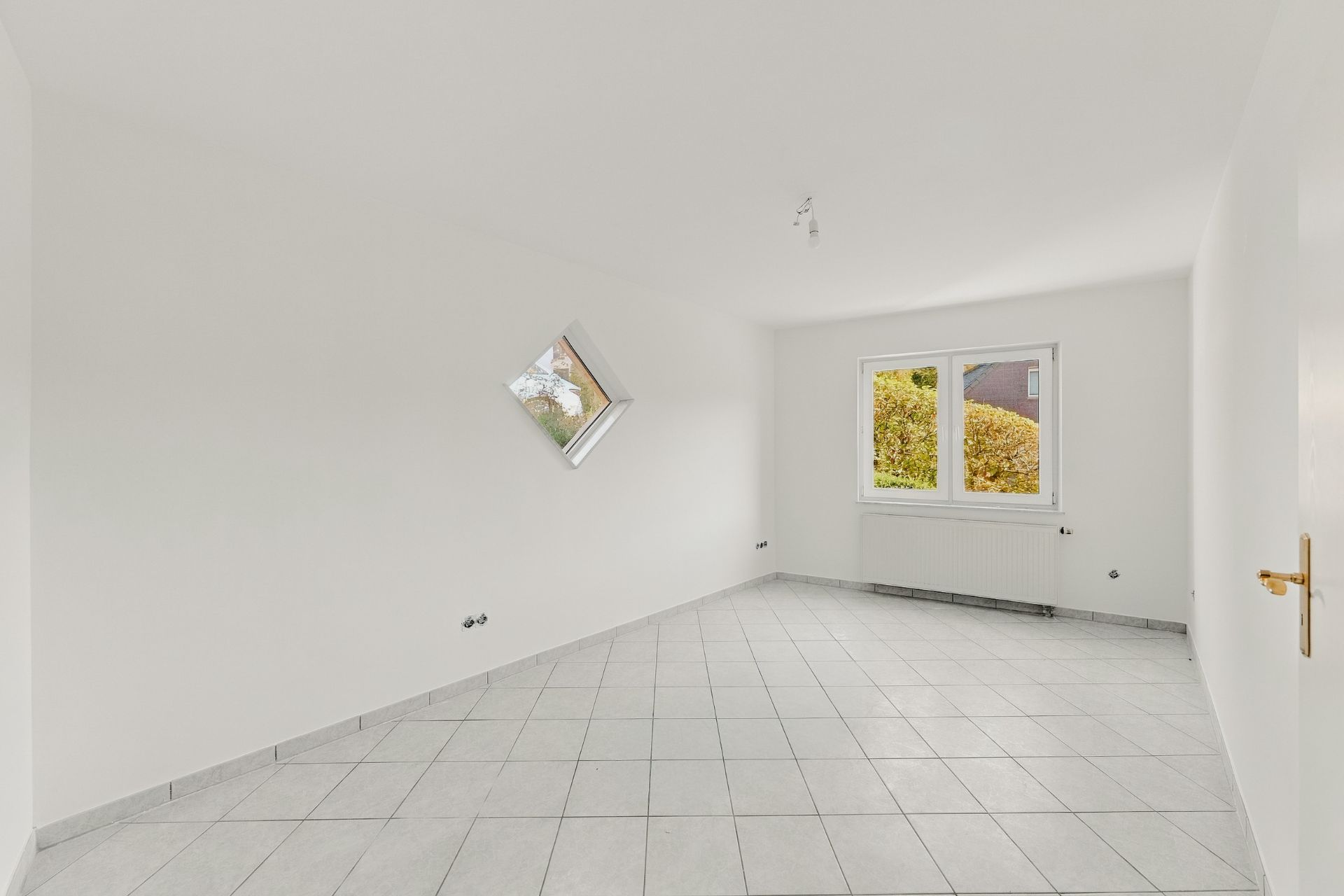 Leerer, weißer Raum mit Fliesenboden und zwei Fenstern, eines quadratisch, eines rechteckig.