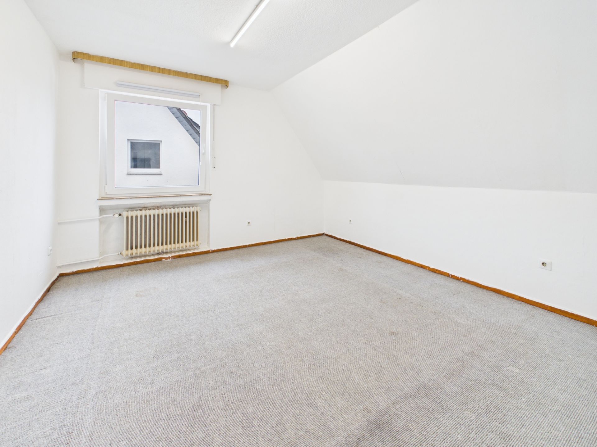 Leeres Zimmer mit Teppichboden, weiß gestrichenen Wänden und einem Fenster mit Heizkörper darunter.