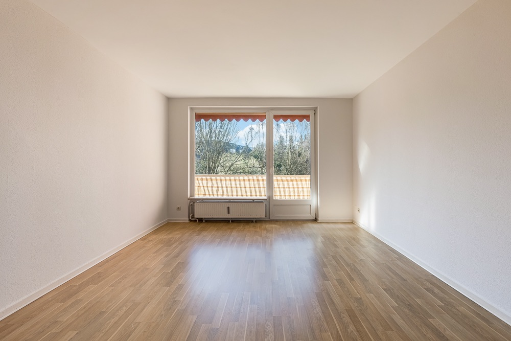 Leerer Raum mit Holzfußboden und großem Fenster, Blick auf Bäume und Berge.