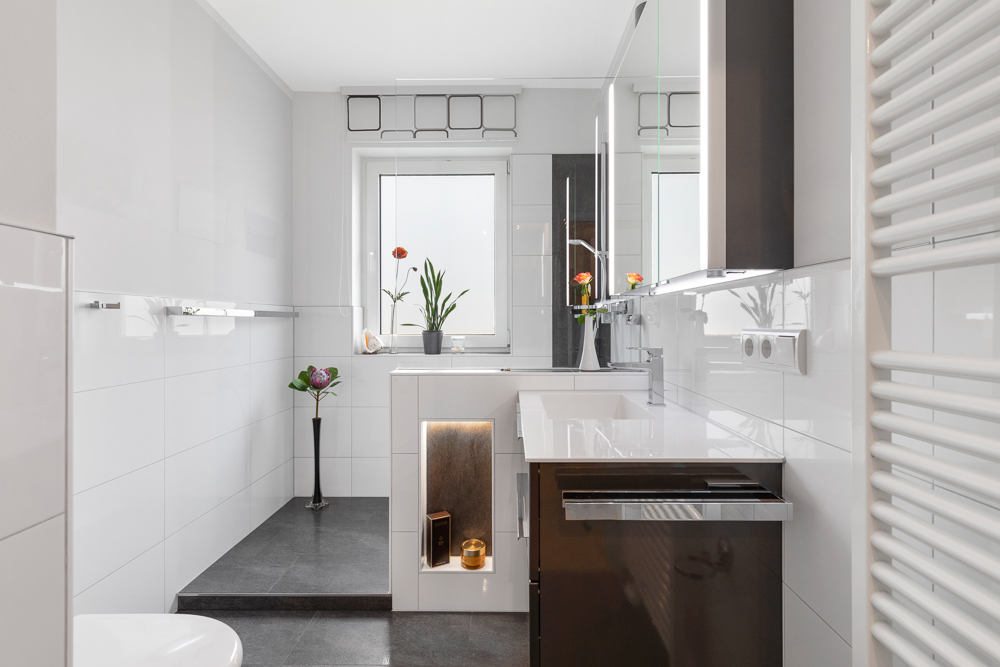 Modernes Badezimmer mit weißem Waschbecken, Spiegel, Fenster und dekorativen Pflanzen.