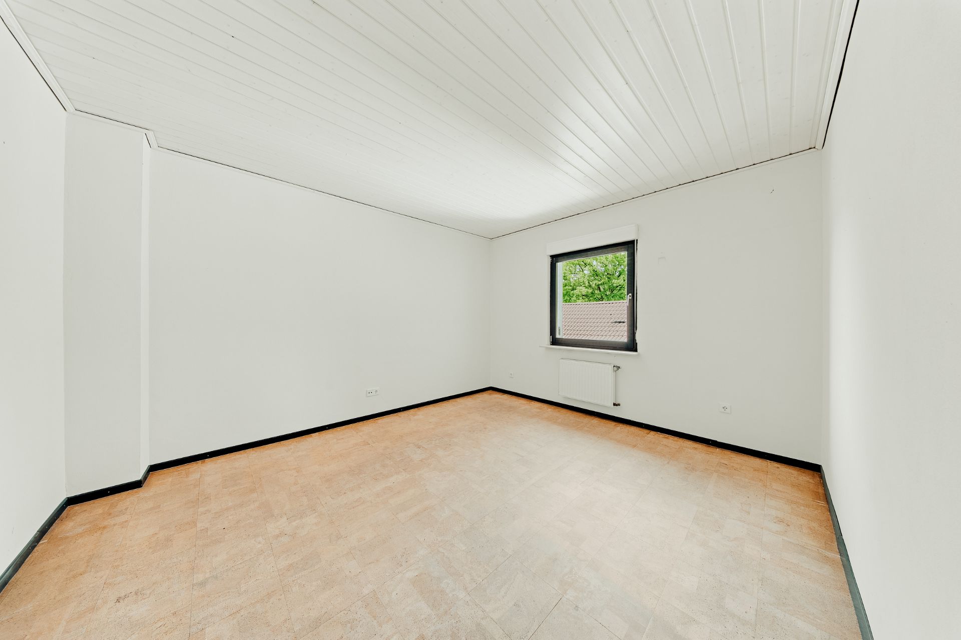 Leeres Zimmer mit weiß gestrichenen Wänden, Holzfußboden und einem Fenster mit Blick ins Grüne.