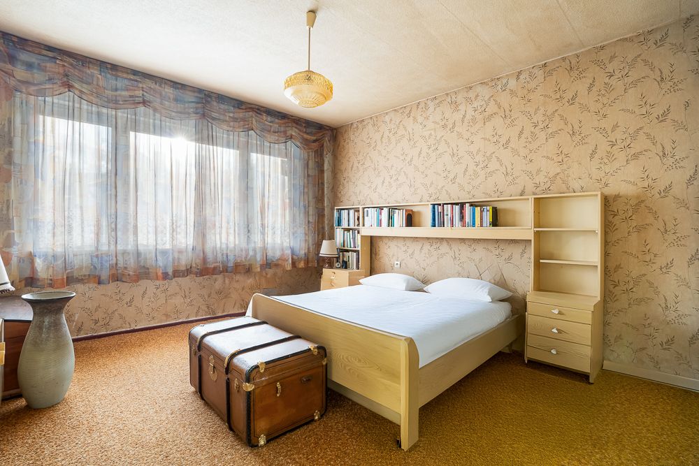 Schlafzimmer mit Doppelbett, Bücherregal im Kopfteil, zwei Koffern und großen Fenstern mit Gardinen.