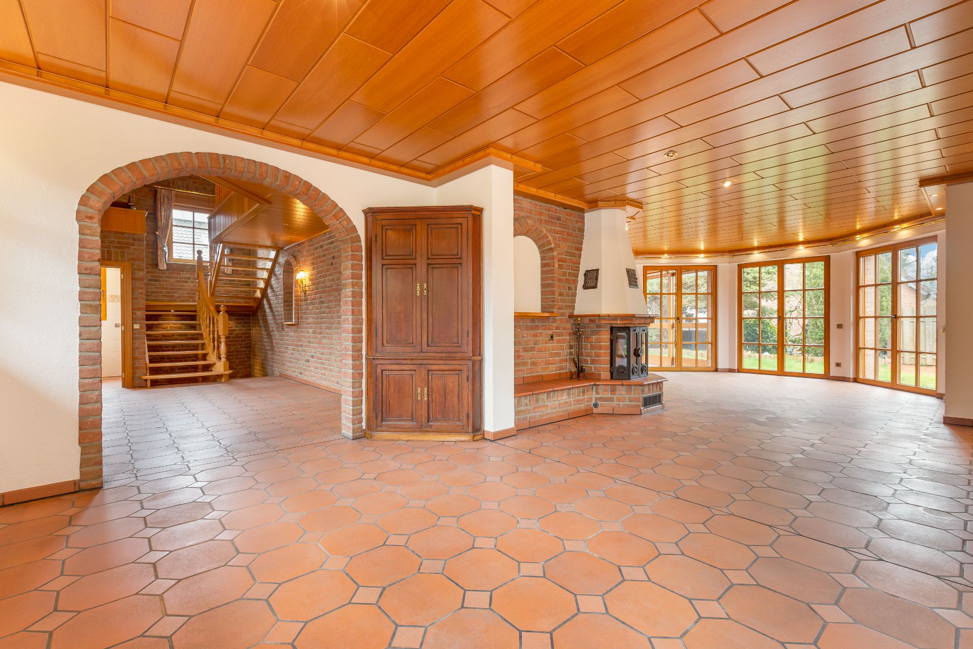 Heller Wohnraum mit Fliesenboden, Holzdecke, Kamin, großen Fenstern und offener Treppe.