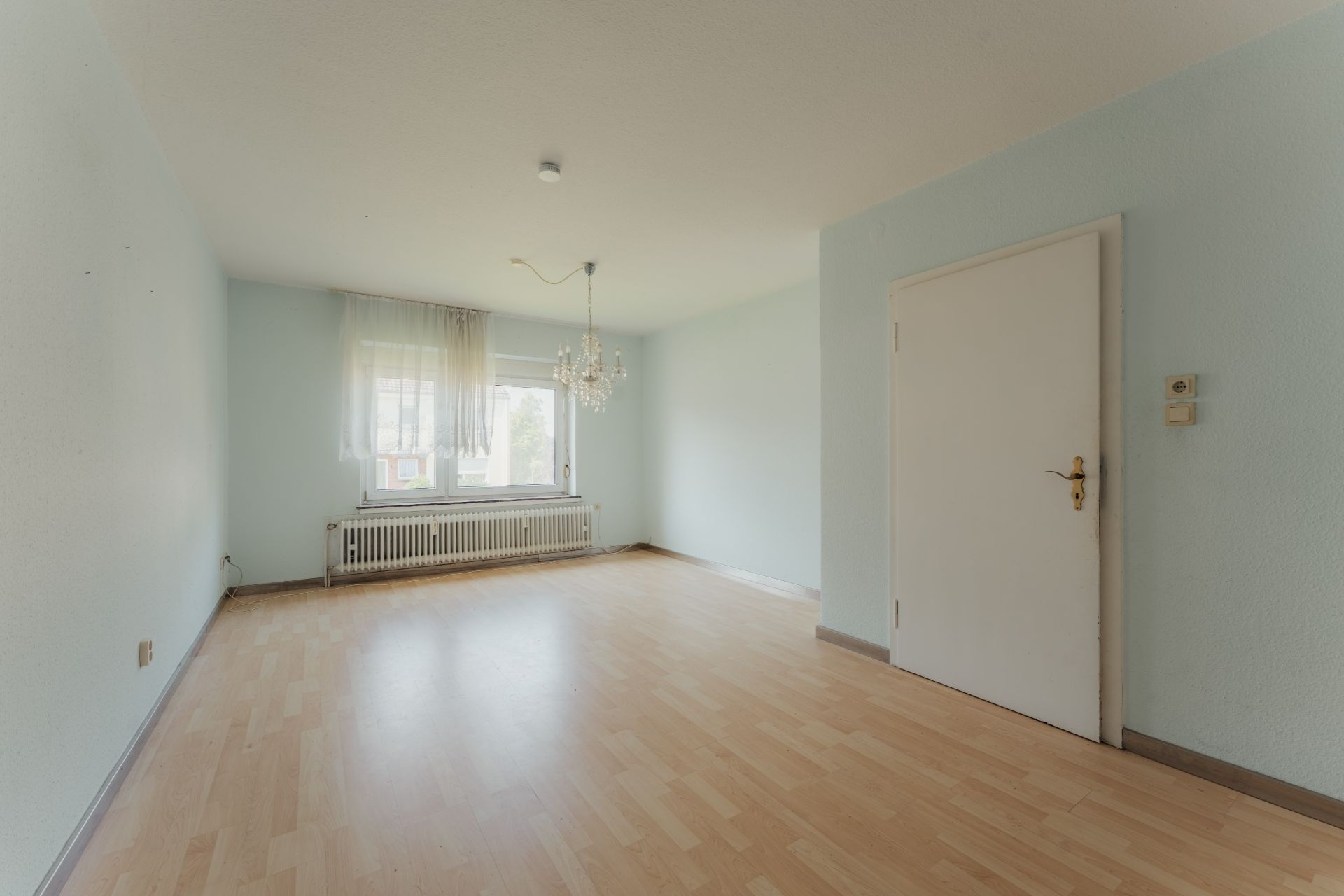 Leeres Zimmer mit Holzfußboden, großem Fenster, Heizkörper und Kronleuchter.