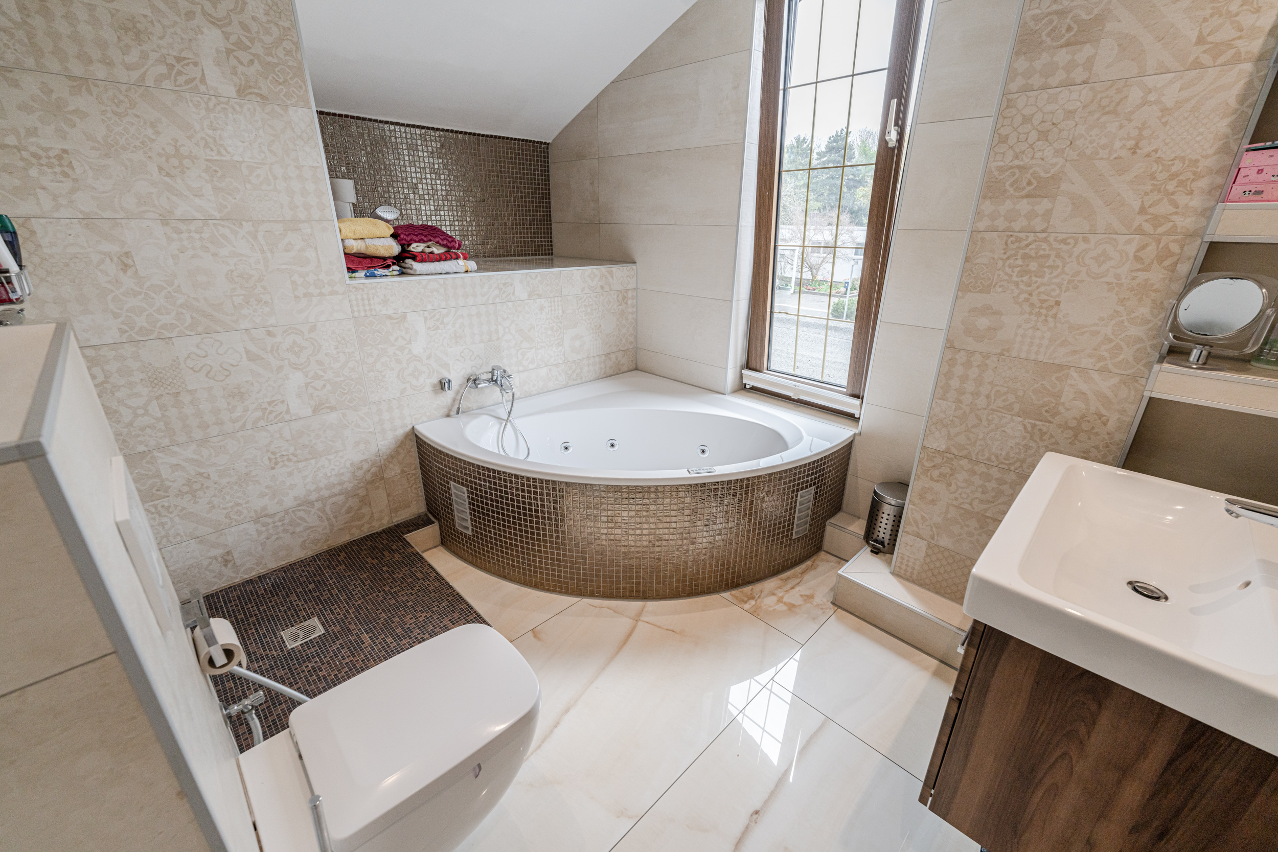 Modernes Badezimmer mit Eckbadewanne, Toilette und Fenster. Handtücher liegen auf einem Regal.