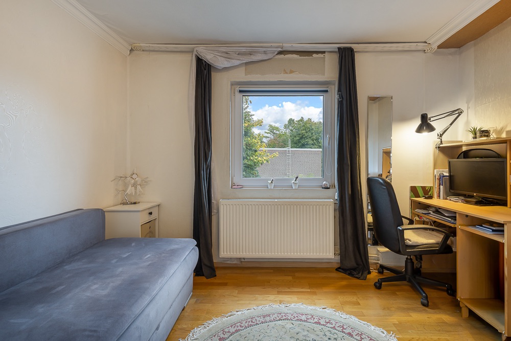 Helles Zimmer mit grauem Sofa, Schreibtisch mit Computer, Stuhl und Fenster mit Vorhängen.