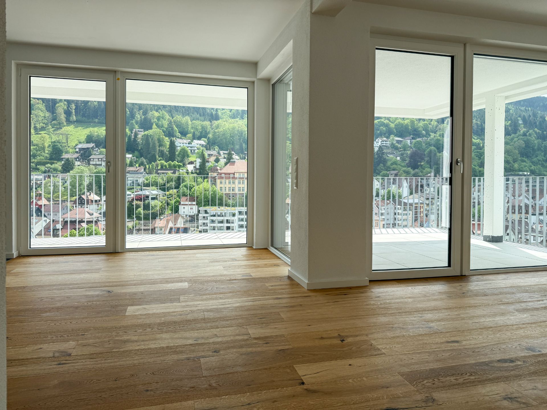 Helles Wohnzimmer mit Holzfußboden und großen Fenstern, Blick auf einen Balkon und grüne Hügel.