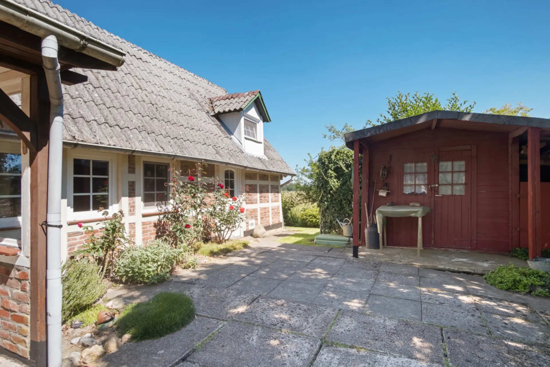 Innenhof mit Blumenbeeten, Pflastersteinen, einem roten Gartenschuppen und einem Haus mit Ziegeldach.