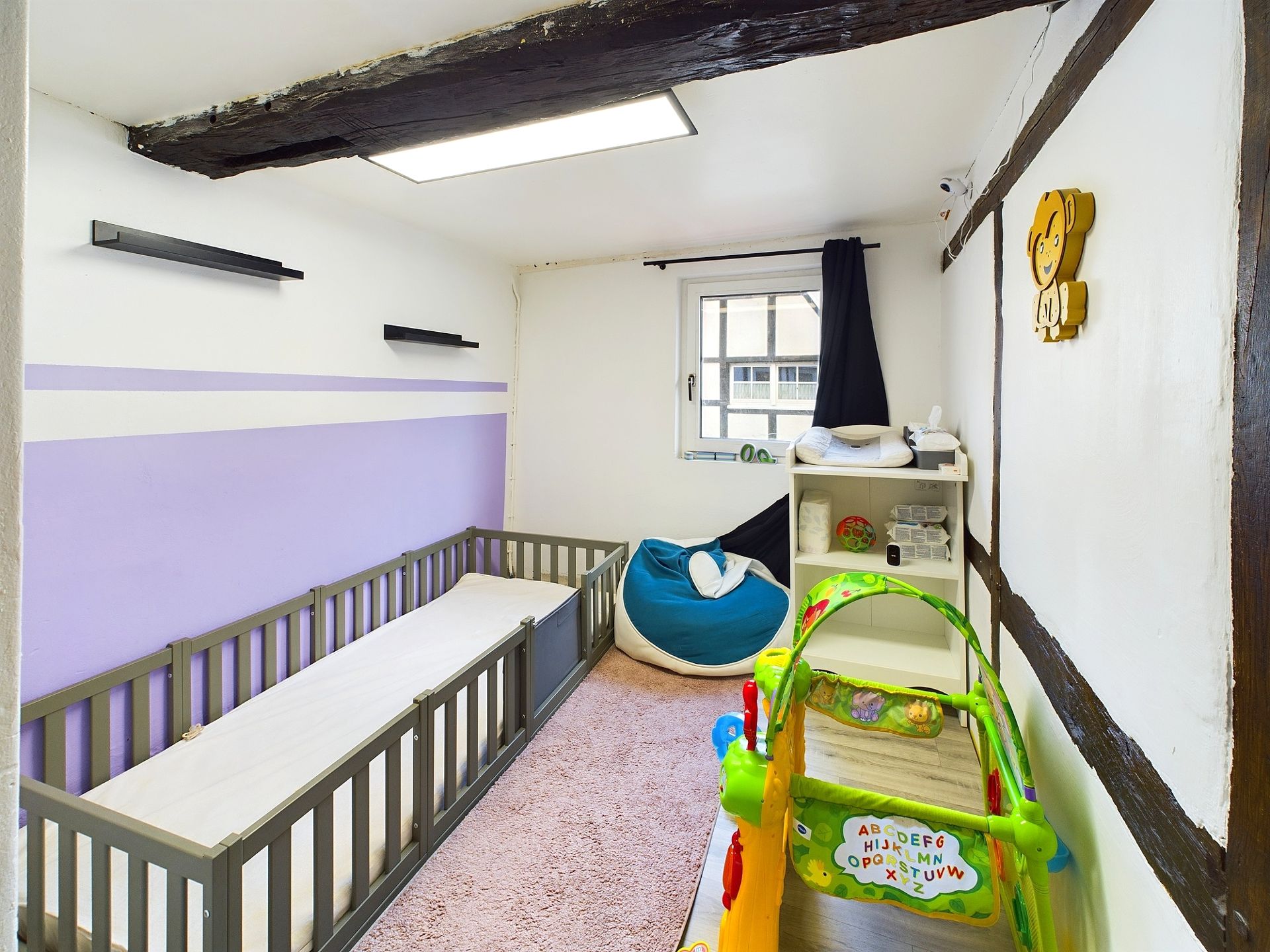 Kinderzimmer mit Gitterbett, Spielzeug, Regal und Fenster, Holzbalken an der Decke.
