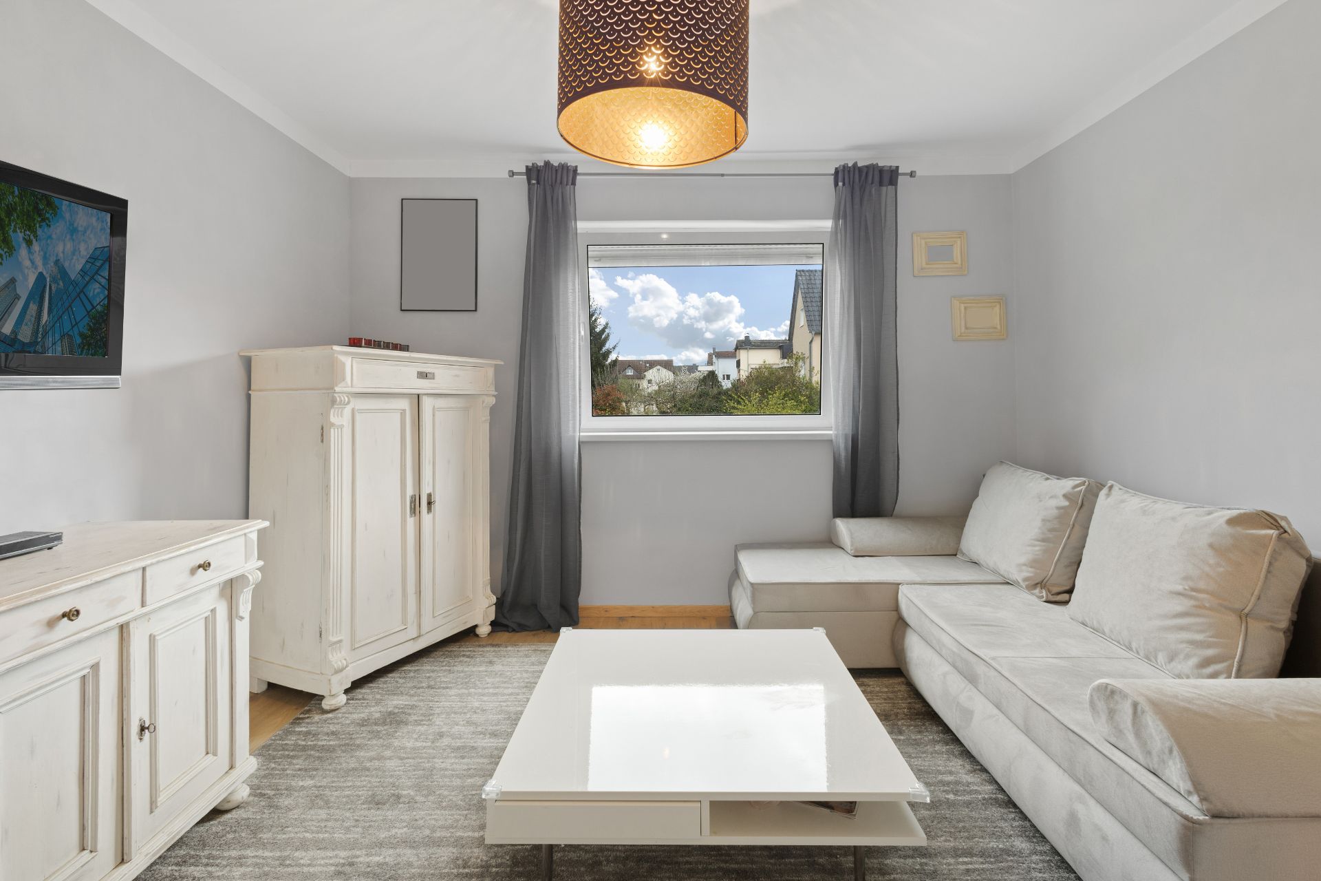 Helles Wohnzimmer mit grauem Sofa, Couchtisch, Schrank, TV und großem Fenster mit Blick nach draußen.