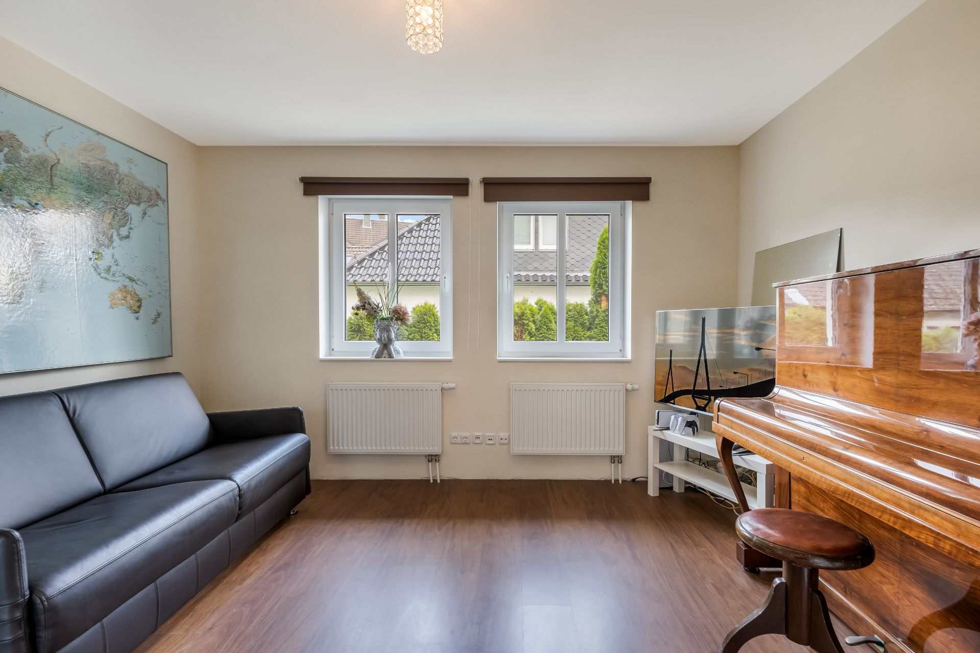 Helles Wohnzimmer mit Sofa, Klavier, Weltkarte an der Wand und zwei Fenstern.