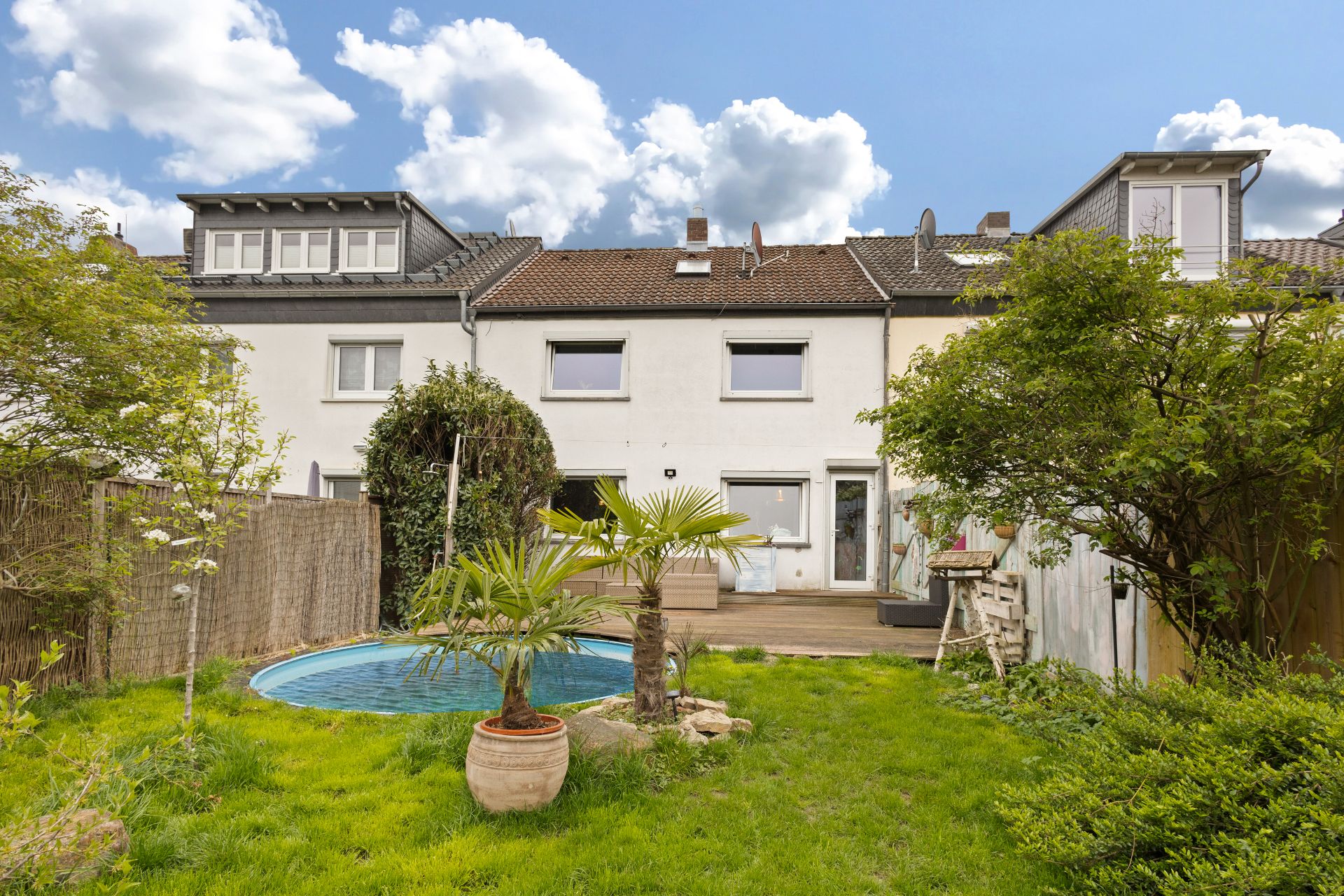 Rückseite eines Reihenhauses mit Garten, Terrasse, Pool und grüner Bepflanzung.