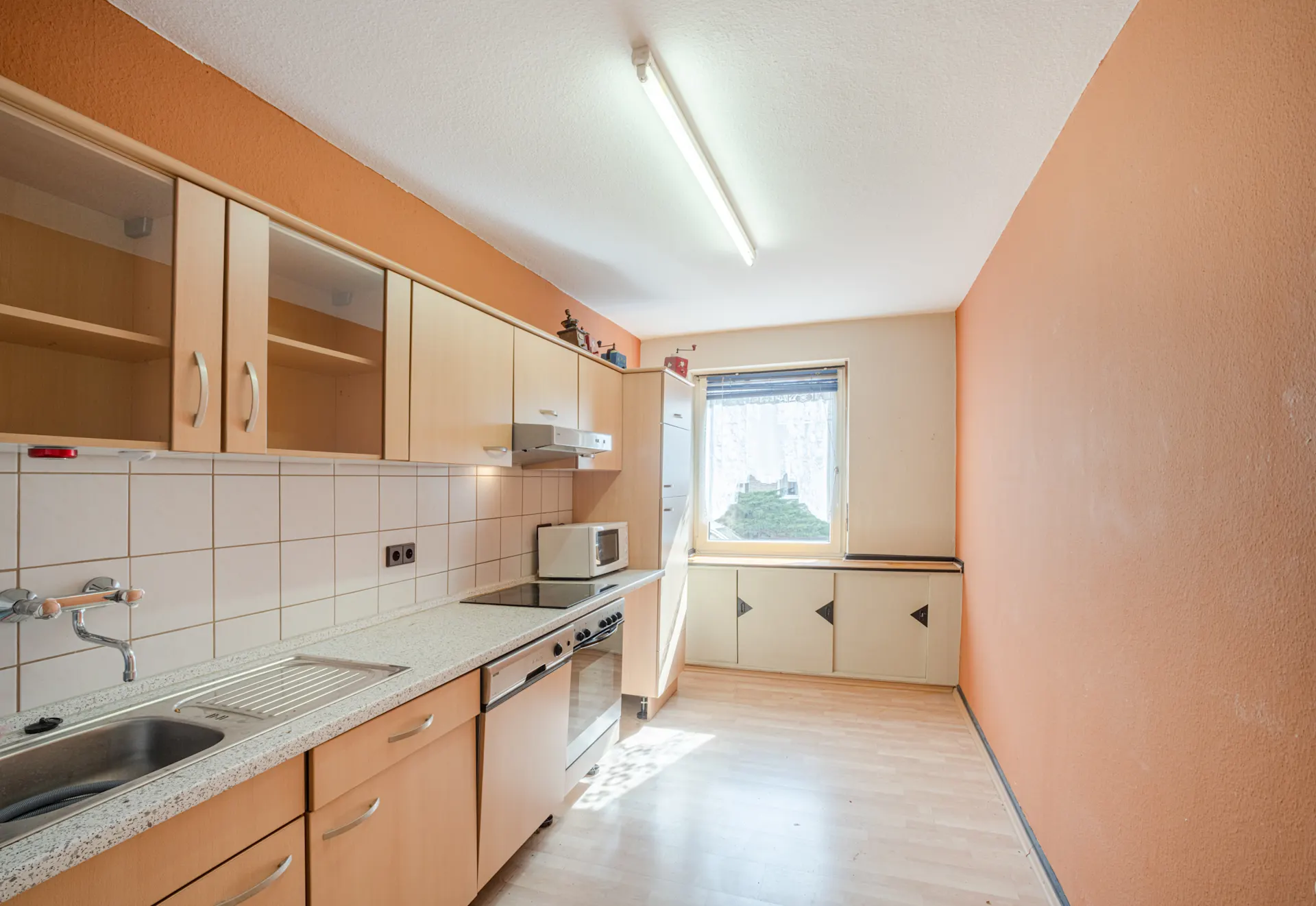 Lange Küche mit hellen Schränken, Elektroherd, Spüle und Fenster mit Blick ins Freie.