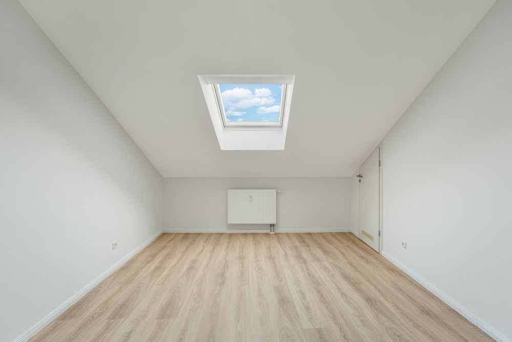 Leerer, heller Raum mit Dachfenster, weißem Heizkörper und hellem Holzboden.