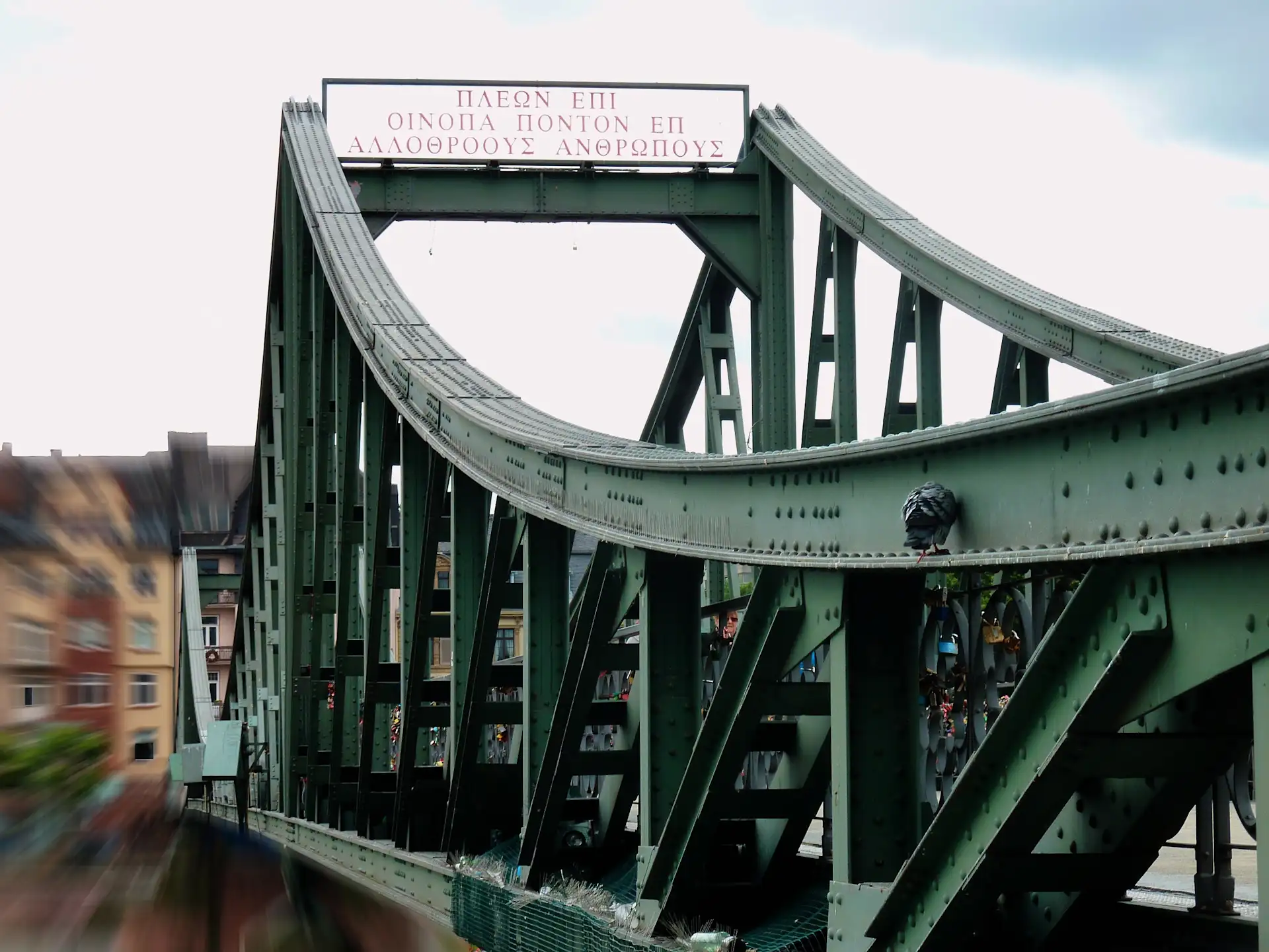 Grüne Metallbrücke mit griechischem Schriftzug, im Hintergrund Wohnhäuser.