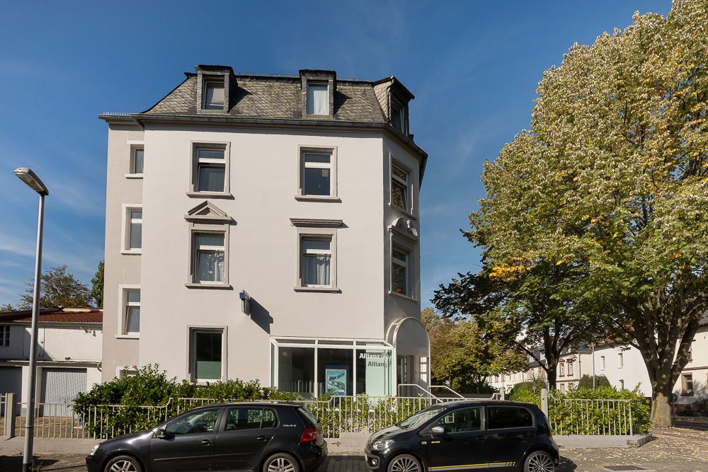 Mehrstöckiges Wohnhaus mit grauem Dach und mehreren Fenstern, davor stehen zwei Autos auf der Straße.