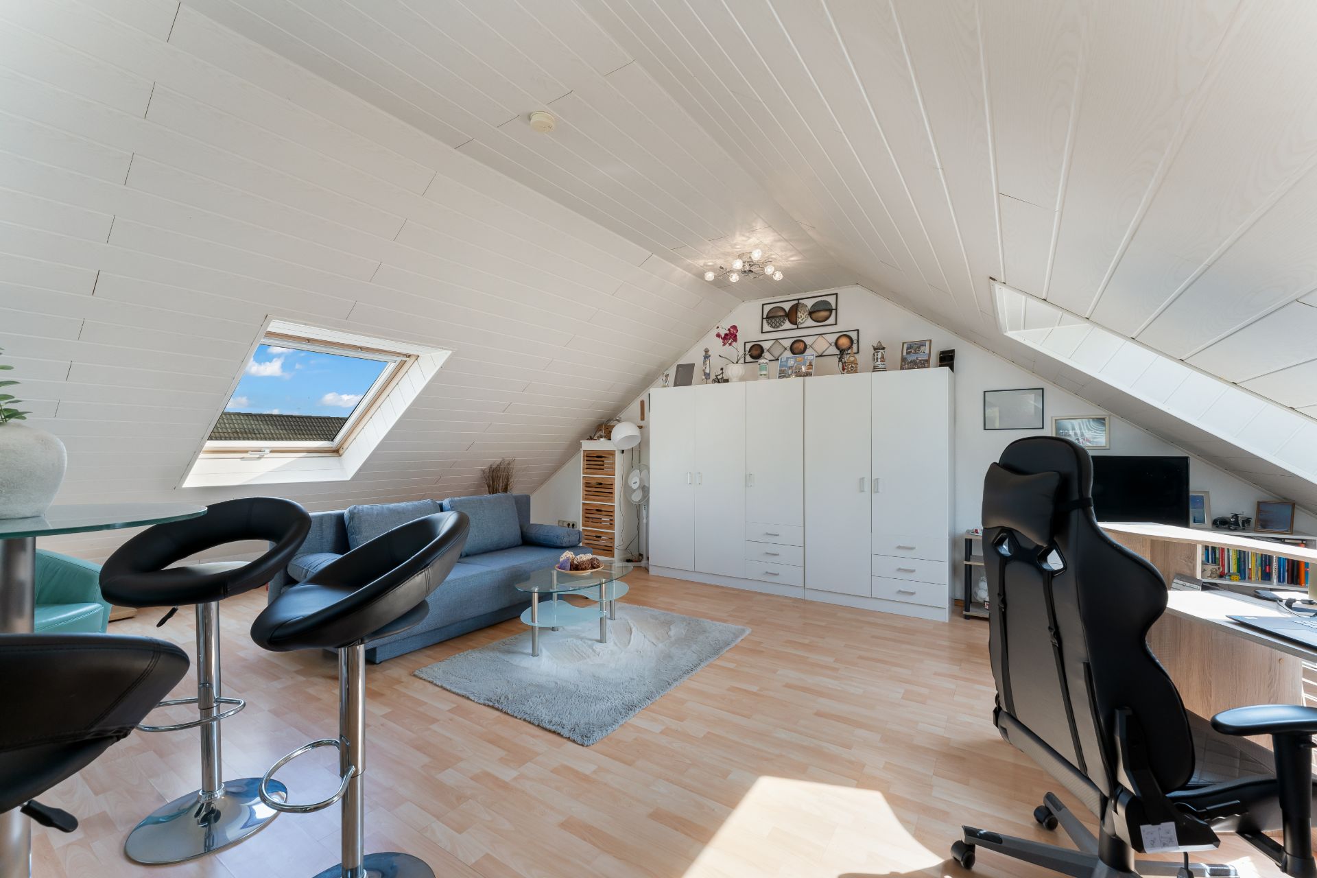 Helles Dachzimmer mit Sofa, Schreibtisch, Barhockern und weißen Einbauschränken.