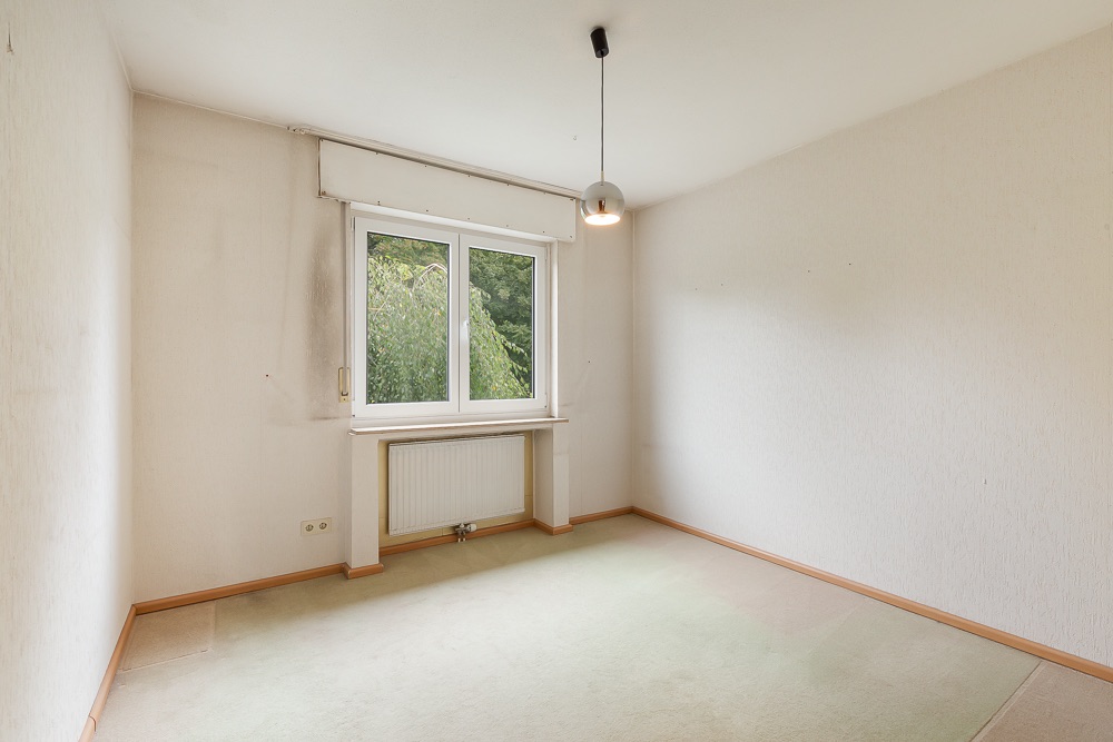 Leeres Zimmer mit Teppichboden, großem Fenster und Heizkörper darunter.